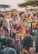 A group of people adorned in colorful traditional attire, featuring intricate beadwork and patterns. The individuals appear engaged in conversation and smiling, with a woman carrying a child on her back. The background includes sparse trees, suggesting a rural setting.