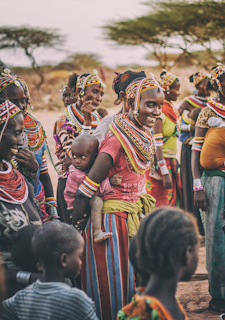A group of people adorned in colorful traditional attire, featuring intricate beadwork and patterns. The individuals appear engaged in conversation and smiling, with a woman carrying a child on her back. The background includes sparse trees, suggesting a rural setting.