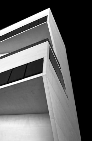 A modern, minimalist architectural structure with angular lines and large, dark windows. It is composed of white concrete and is set against a stark, black background, enhancing its geometric design.