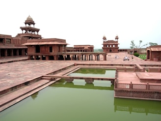 A historical architectural site featuring red sandstone buildings with intricate carvings and multiple storeys. There is a large courtyard area with a rectangular water tank filled with greenish water. A few people can be seen walking in the background, suggesting this is a tourist site.