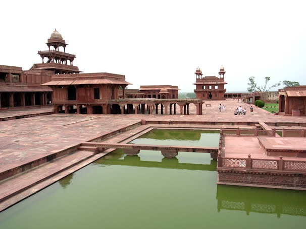 A historical architectural site featuring red sandstone buildings with intricate carvings and multiple storeys. There is a large courtyard area with a rectangular water tank filled with greenish water. A few people can be seen walking in the background, suggesting this is a tourist site.