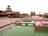 A historical architectural site featuring red sandstone buildings with intricate carvings and multiple storeys. There is a large courtyard area with a rectangular water tank filled with greenish water. A few people can be seen walking in the background, suggesting this is a tourist site.