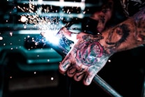 A close-up of a tattooed hand holding a welding torch. Sparks are flying from the welding process and the focus is on the intensity of the action. The background is blurred, drawing attention to the detailed tattoos and the bright welding sparks.