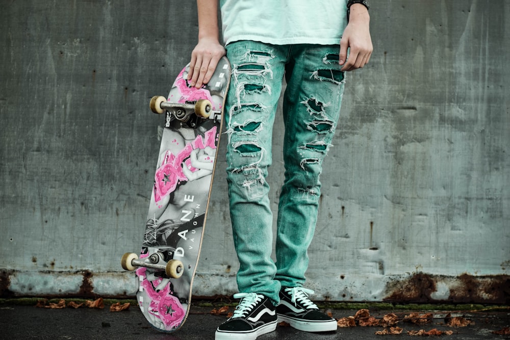 Mann steht neben der Wand und hält ein graues und rosa Skateboard in der Hand
