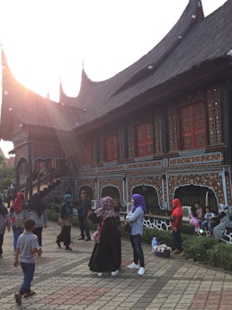 A group of people are gathered in front of a traditional building with detailed, ornate patterns. The building has a distinctive roof design and intricate wooden decorations. Some children and adults are sitting or standing nearby, engaging in casual conversation. The sun is setting, casting a warm glow over the scene.