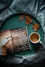 An open book with inspirational text is placed on a teal tray alongside a white cup of coffee and a few dried brown oak leaves. The background features a textured fabric in a deep teal color.