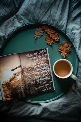 An open book with inspirational text is placed on a teal tray alongside a white cup of coffee and a few dried brown oak leaves. The background features a textured fabric in a deep teal color.