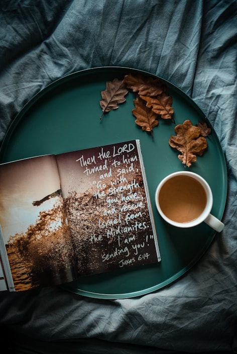 An open book with inspirational text is placed on a teal tray alongside a white cup of coffee and a few dried brown oak leaves. The background features a textured fabric in a deep teal color.