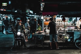 A bustling night market scene with several people interacting around various food stalls. One person rides a motorcycle in the foreground, while others are engaged in buying or selling food items under bright artificial lights. The area is lively with a mix of modern and traditional elements.