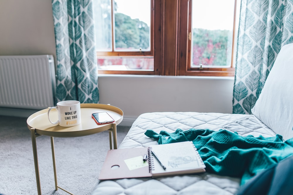 Colchón blanco acolchado con cuaderno de espiral y edredón verde junto a una mesa auxiliar redonda amarilla dentro de la habitación