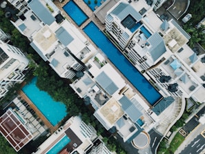 Aerial view of a residential complex featuring multiple swimming pools situated between modern apartment buildings. The architecture includes clean lines and flat roofs, with ample greenery surrounding the area. Loungers and poolside furniture are arranged neatly, suggesting a luxurious and organized environment.