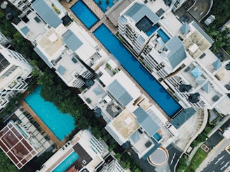 Aerial view of a residential complex featuring multiple swimming pools situated between modern apartment buildings. The architecture includes clean lines and flat roofs, with ample greenery surrounding the area. Loungers and poolside furniture are arranged neatly, suggesting a luxurious and organized environment.