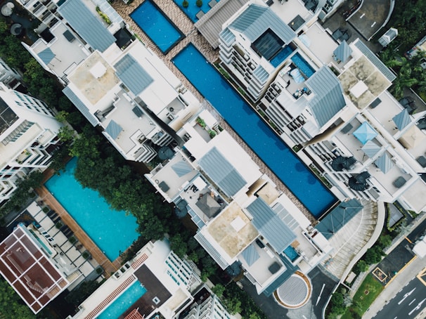 Aerial view of a residential complex featuring multiple swimming pools situated between modern apartment buildings. The architecture includes clean lines and flat roofs, with ample greenery surrounding the area. Loungers and poolside furniture are arranged neatly, suggesting a luxurious and organized environment.