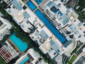 Aerial view of a residential complex featuring multiple swimming pools situated between modern apartment buildings. The architecture includes clean lines and flat roofs, with ample greenery surrounding the area. Loungers and poolside furniture are arranged neatly, suggesting a luxurious and organized environment.