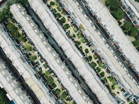 An aerial view of a residential area featuring rows of identical houses with gabled roofs. The houses are aligned in parallel, separated by narrow pathways and interspersed with small green spaces containing trees and shrubs.