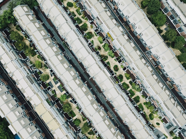 An aerial view of a residential area featuring rows of identical houses with gabled roofs. The houses are aligned in parallel, separated by narrow pathways and interspersed with small green spaces containing trees and shrubs.