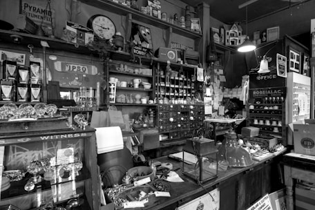An old-fashioned shop interior with various items on display, including vintage bottles, drawers, shelves filled with knick-knacks, and a hanging lamp. The setting is cluttered but organized, with an assortment of products and handwritten notes pinned to the wall.