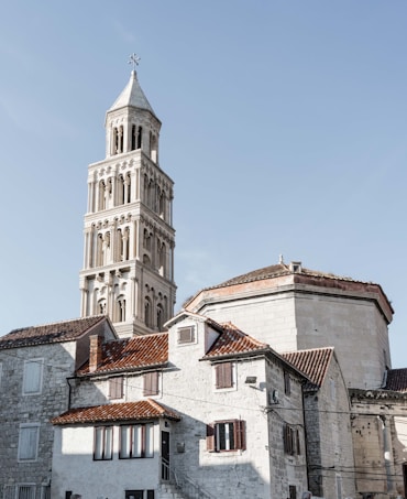 A stone tower with intricate architectural details rises prominently against a clear blue sky. The foreground features historic stone buildings with red-tiled roofs and wooden shutters. The scene conveys a sense of historical and architectural significance.