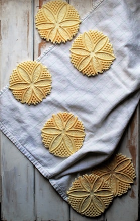 Round, intricately patterned cookies are arranged on a white cloth that rests on a wooden surface. The cookies have a floral, embossed design that gives them a decorative appearance. The cloth is slightly wrinkled, creating a casual and homey setting.