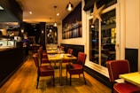 A warmly lit restaurant interior with wooden flooring and modern decor. Red cushioned chairs surround tables set with glassware and napkins. The space has a cozy atmosphere with hanging pendant lights and a large framed photograph of a cityscape on the wall. A large decorative star hangs near the window.