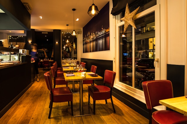 A warmly lit restaurant interior with wooden flooring and modern decor. Red cushioned chairs surround tables set with glassware and napkins. The space has a cozy atmosphere with hanging pendant lights and a large framed photograph of a cityscape on the wall. A large decorative star hangs near the window.
