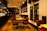 A warmly lit restaurant interior with wooden flooring and modern decor. Red cushioned chairs surround tables set with glassware and napkins. The space has a cozy atmosphere with hanging pendant lights and a large framed photograph of a cityscape on the wall. A large decorative star hangs near the window.