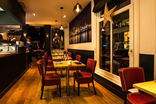 A warmly lit restaurant interior with wooden flooring and modern decor. Red cushioned chairs surround tables set with glassware and napkins. The space has a cozy atmosphere with hanging pendant lights and a large framed photograph of a cityscape on the wall. A large decorative star hangs near the window.
