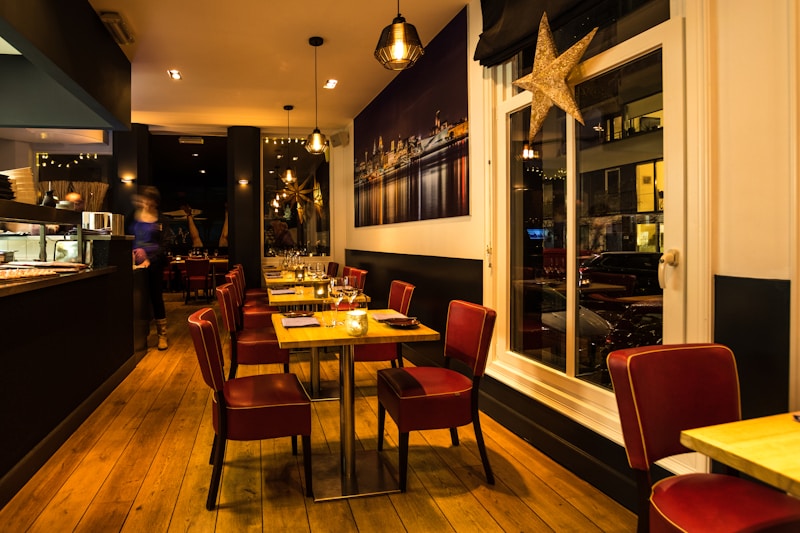 A warmly lit restaurant interior with wooden flooring and modern decor. Red cushioned chairs surround tables set with glassware and napkins. The space has a cozy atmosphere with hanging pendant lights and a large framed photograph of a cityscape on the wall. A large decorative star hangs near the window.