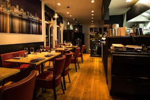 Warmly lit restaurant interior featuring wooden floors and red leather chairs. Tables are set with wine glasses and cutlery, ready for dining. A large photograph of a cityscape at night adorns one wall. The open kitchen area is visible, with cooking equipment and stacks of plates.