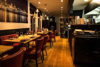 Warmly lit restaurant interior featuring wooden floors and red leather chairs. Tables are set with wine glasses and cutlery, ready for dining. A large photograph of a cityscape at night adorns one wall. The open kitchen area is visible, with cooking equipment and stacks of plates.
