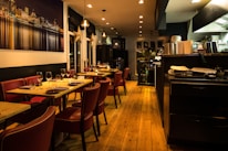 Warmly lit restaurant interior featuring wooden floors and red leather chairs. Tables are set with wine glasses and cutlery, ready for dining. A large photograph of a cityscape at night adorns one wall. The open kitchen area is visible, with cooking equipment and stacks of plates.