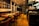Warmly lit restaurant interior featuring wooden floors and red leather chairs. Tables are set with wine glasses and cutlery, ready for dining. A large photograph of a cityscape at night adorns one wall. The open kitchen area is visible, with cooking equipment and stacks of plates.