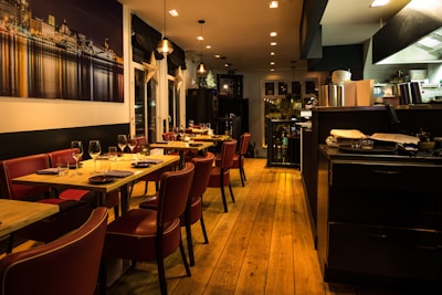 Warmly lit restaurant interior featuring wooden floors and red leather chairs. Tables are set with wine glasses and cutlery, ready for dining. A large photograph of a cityscape at night adorns one wall. The open kitchen area is visible, with cooking equipment and stacks of plates.
