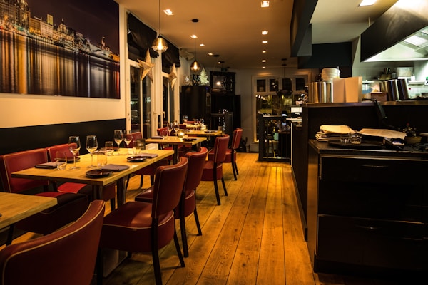 Warmly lit restaurant interior featuring wooden floors and red leather chairs. Tables are set with wine glasses and cutlery, ready for dining. A large photograph of a cityscape at night adorns one wall. The open kitchen area is visible, with cooking equipment and stacks of plates.