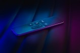 A sleek, modern remote control rests on a dark surface, illuminated by a moody mix of blue and purple lighting. The buttons are prominently displayed on the upper surface, featuring symbols for volume and navigation.