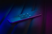 A sleek, modern remote control rests on a dark surface, illuminated by a moody mix of blue and purple lighting. The buttons are prominently displayed on the upper surface, featuring symbols for volume and navigation.