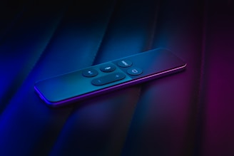 A sleek, modern remote control rests on a dark surface, illuminated by a moody mix of blue and purple lighting. The buttons are prominently displayed on the upper surface, featuring symbols for volume and navigation.