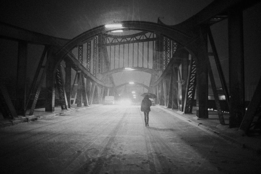 grayscale photo of woman under umbrella walking on suspension bridge