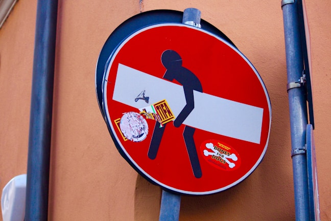 A red circular no-entry road sign has been modified with artistic stickers. A black silhouette figure appears to be carrying the horizontal white bar of the sign. Additional stickers, including one with the text 'BONES FOR DOGS', add to the playful alteration.