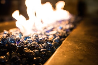 A close-up view of blue glass stones with flames rising above them, creating a glowing effect. The focus is on the shimmering texture of the stones, with the fire adding a dynamic and warm element.