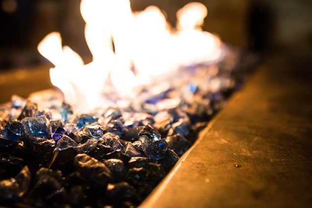A close-up view of blue glass stones with flames rising above them, creating a glowing effect. The focus is on the shimmering texture of the stones, with the fire adding a dynamic and warm element.