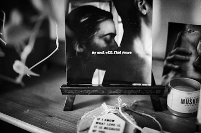 A black and white photograph is displayed on a small easel. The image captures an intimate moment between two people, and text overlay reads 'my soul will find yours.' Surrounding the photo are various objects, including a candle and tags, creating a cozy atmosphere.