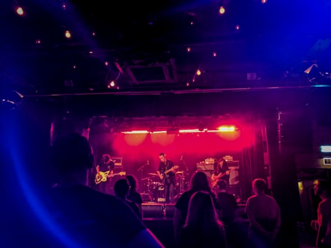 A band performs on a dimly lit stage with vibrant pink and blue lights creating a moody atmosphere. Several people in the audience are visible in the foreground, silhouetted by the lighting.