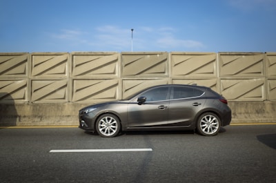 A gray sedan is parked on a freeway next to a large beige concrete barrier. The sky is clear and blue.