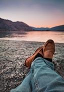 person wearing brown loafers sitting beside body of water