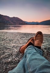 person wearing brown loafers sitting beside body of water