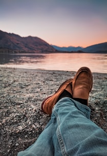 person wearing brown loafers sitting beside body of water