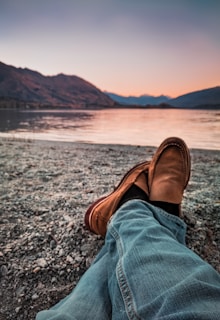 person wearing brown loafers sitting beside body of water