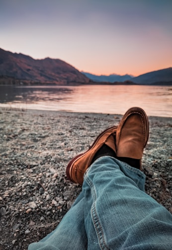 person wearing brown loafers sitting beside body of water