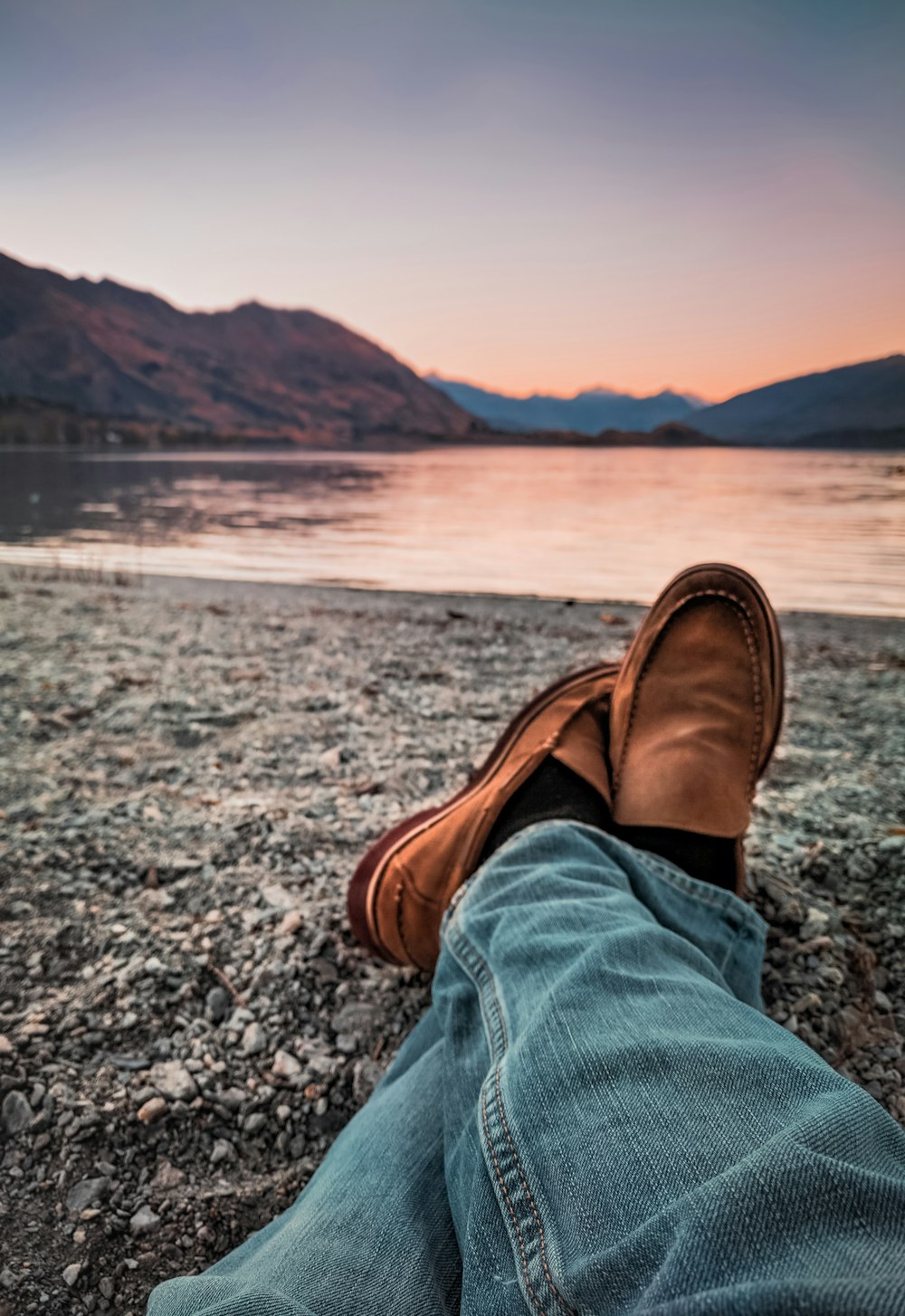 person wearing brown loafers sitting beside body of water